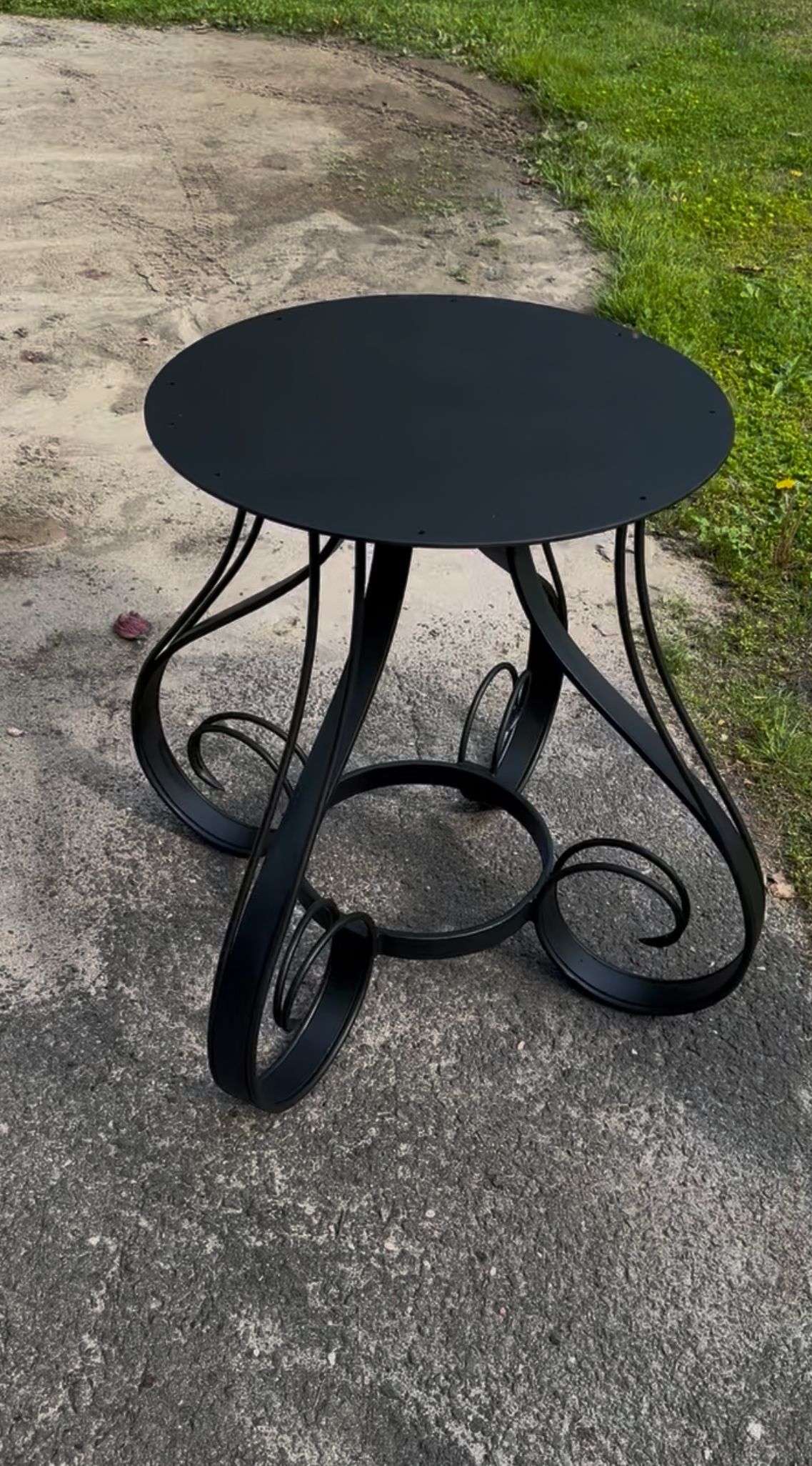 A black table is sitting on a sidewalk next to a grassy field.