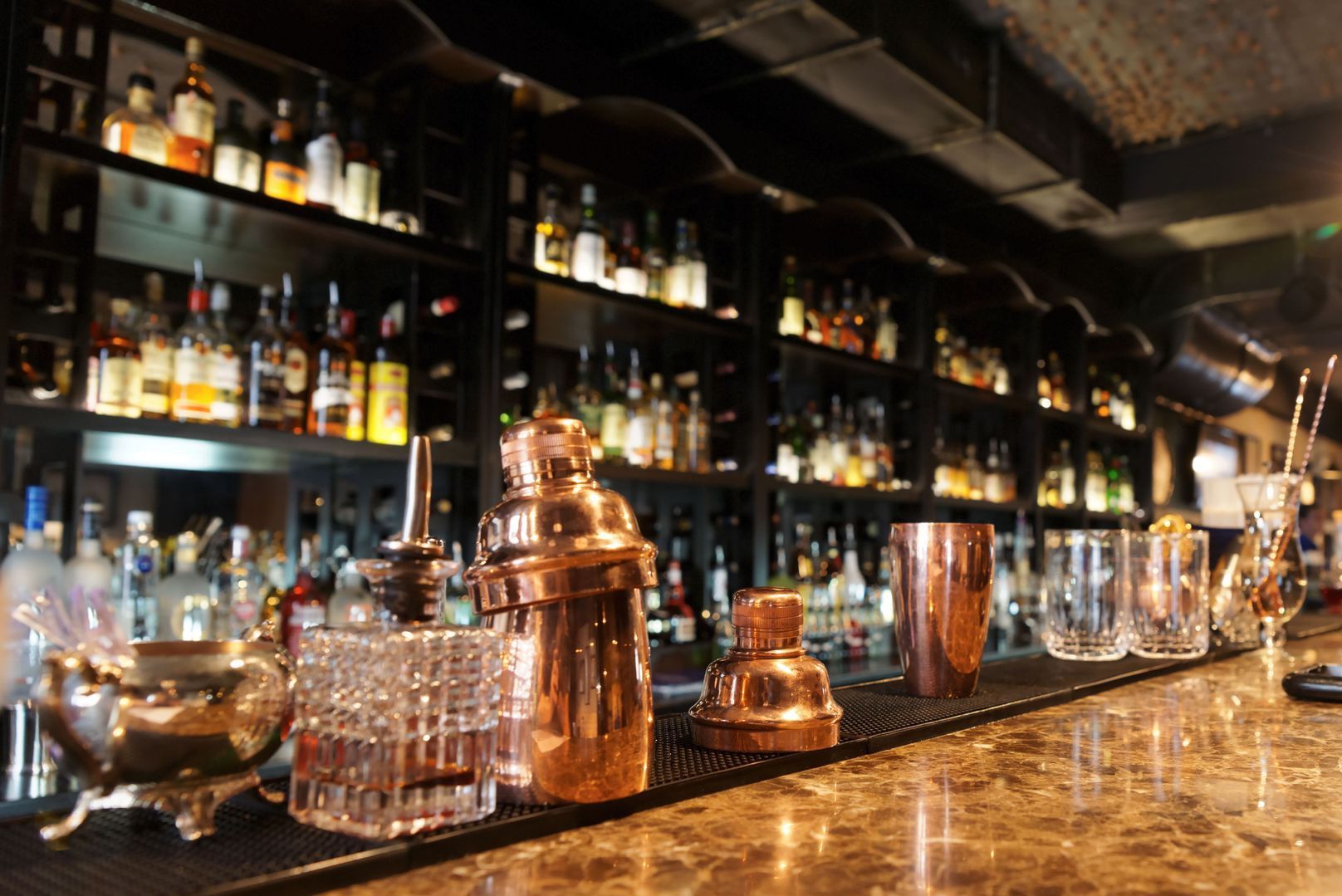 Bar interior with copper cocktail tools on counter, bottles of liquor on shelves in background.