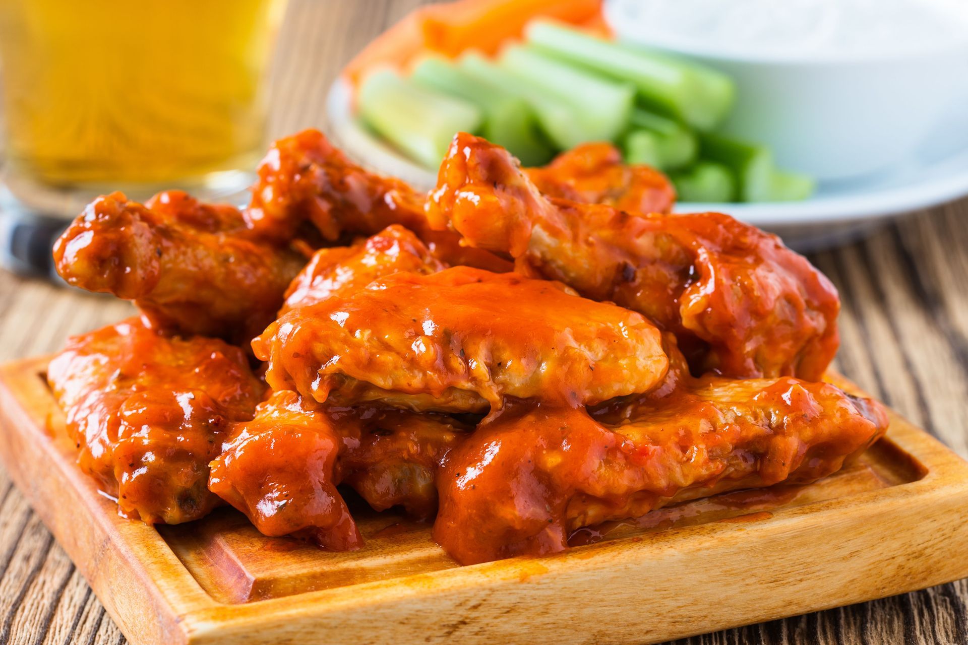 Pile of buffalo wings on a wooden board, with vegetables, dipping sauce, and a beer in the background.