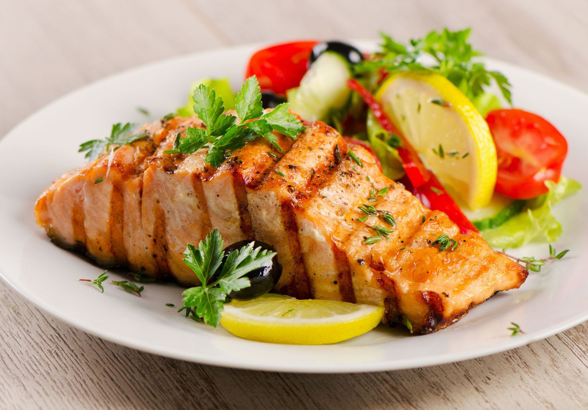 Grilled salmon fillet on a white plate with a side salad of tomatoes, cucumber, olives, and lemon.