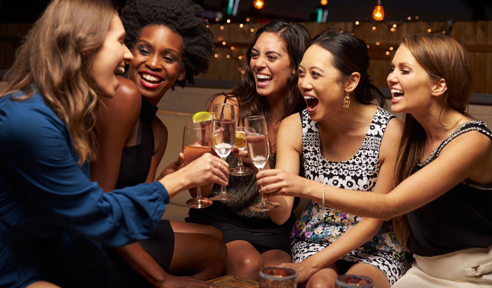 Five diverse women laughing and toasting with champagne flutes in a dimly lit setting.