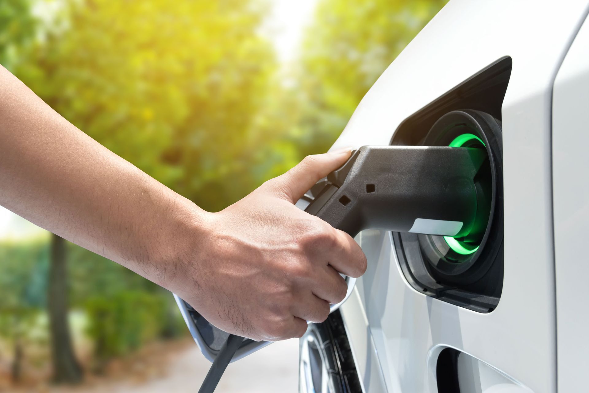 Hand plugging a charging cable into an electric car's port, outdoors. Green light glows.