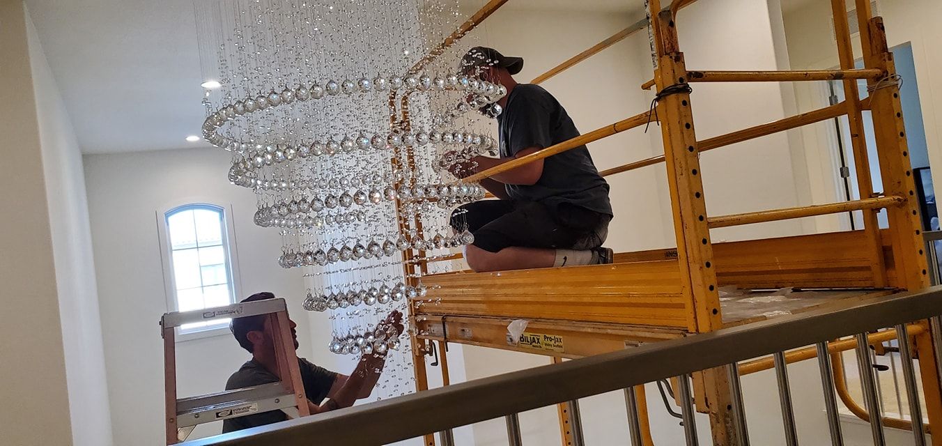 Two workers installing a crystal chandelier, using a scaffold in a building's stairwell.