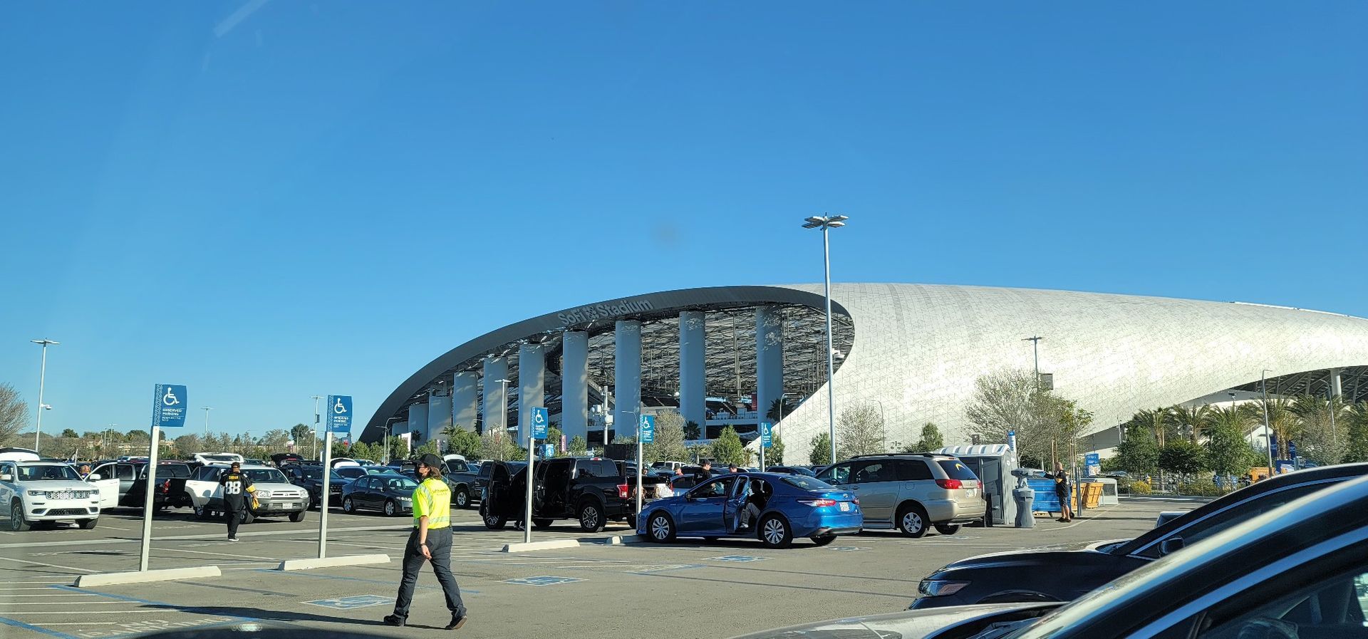 Exterior view of a stadium with a parking lot. A person walks near parked cars under a clear, blue sky.