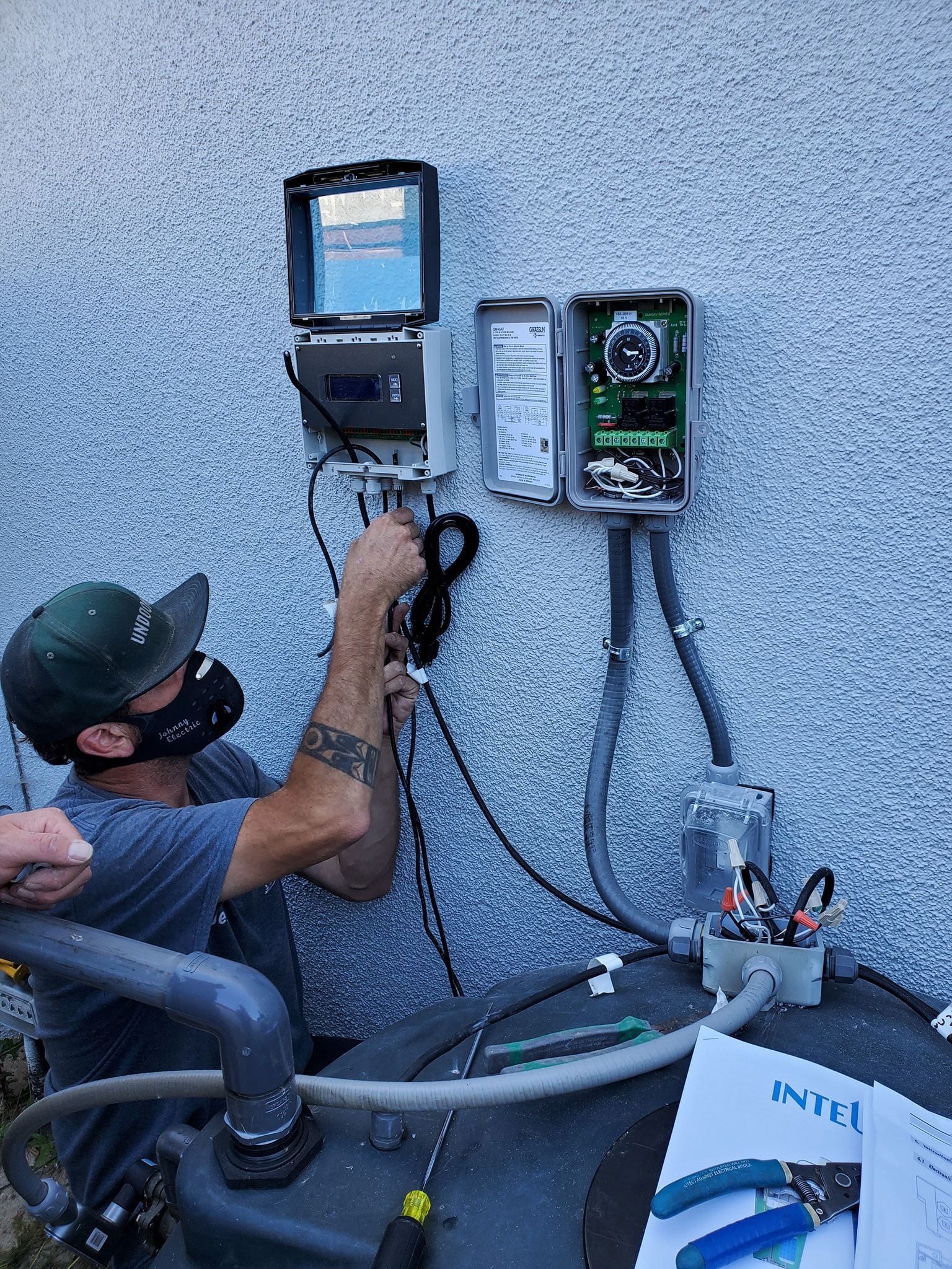 Person in mask connecting wires to a control box mounted on a stucco wall, near a water tank.