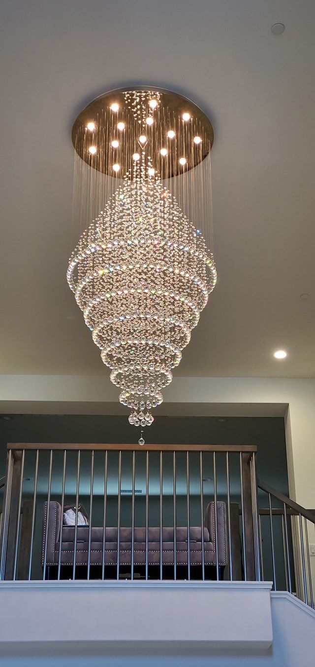 A large, ornate crystal chandelier hangs above a railing and a sofa, in a brightly lit room.
