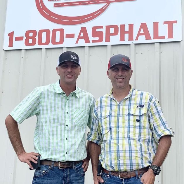 Two men in plaid shirts and baseball caps stand in front of a white wall with a 1-800-ASPHALT sign.