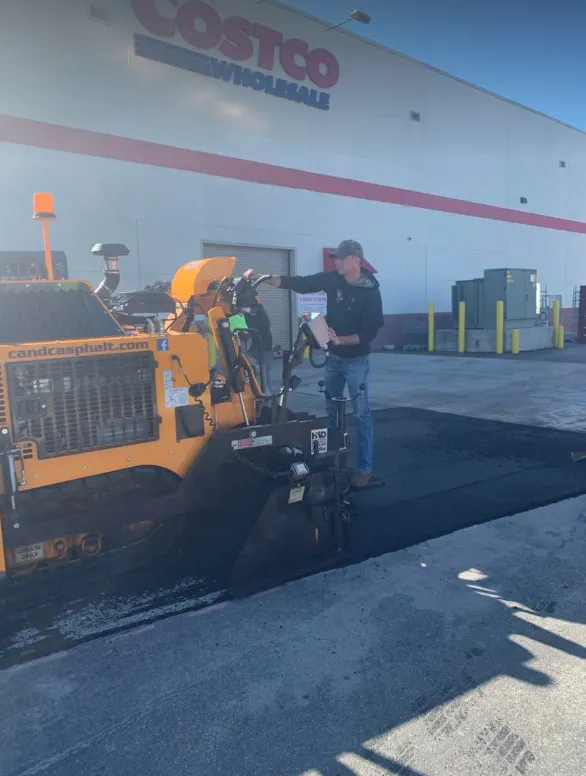 A person operates a yellow paving machine to lay fresh asphalt in front of a Costco Wholesale building.