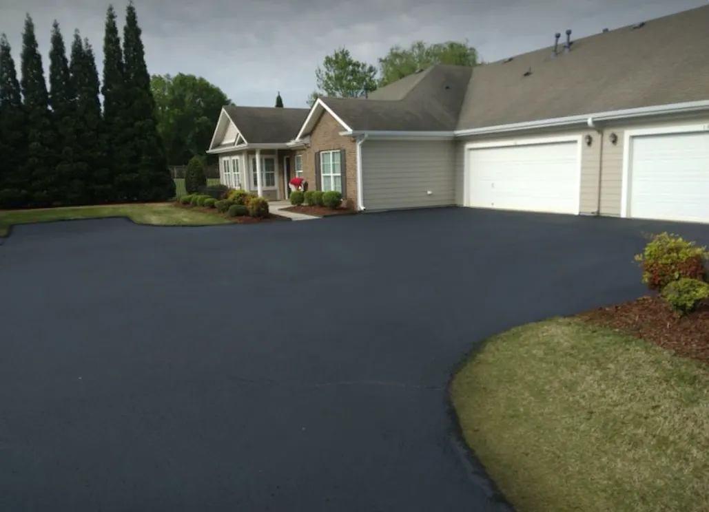 A freshly paved, smooth dark asphalt driveway leading to a multi-unit suburban house with two closed garage doors.