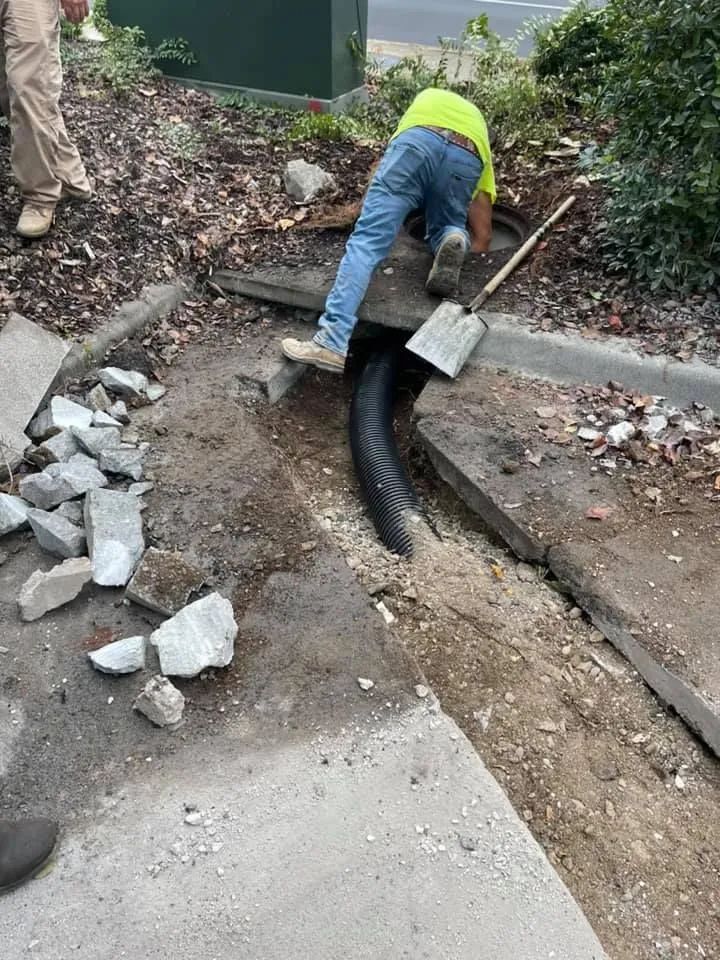 A worker in a yellow shirt reaches into an open storm drain, with a shovel resting nearby in a dirt-filled roadside area.