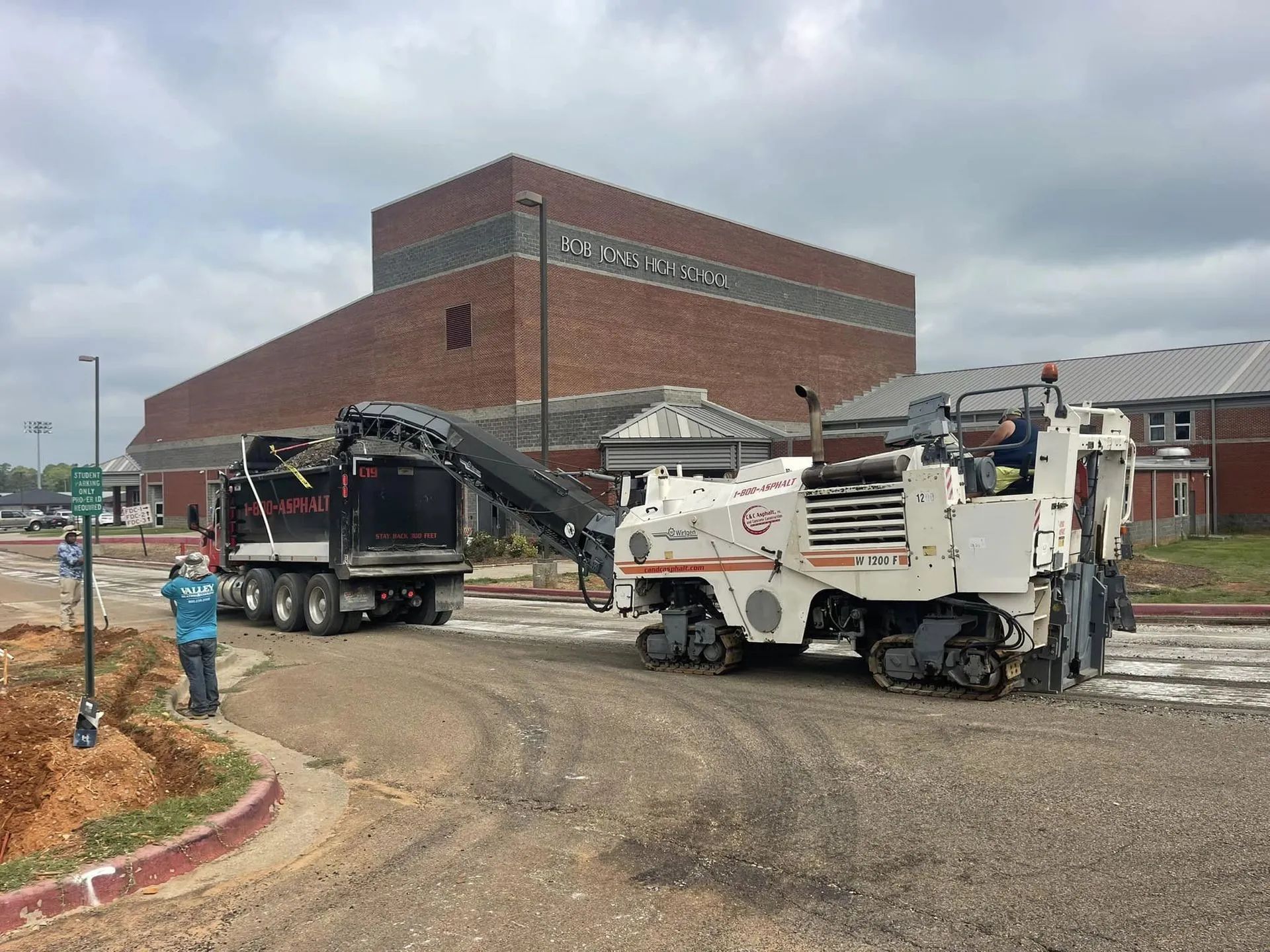 A white road milling machine loads debris into a dump truck in front of a brick building on an overcast day.