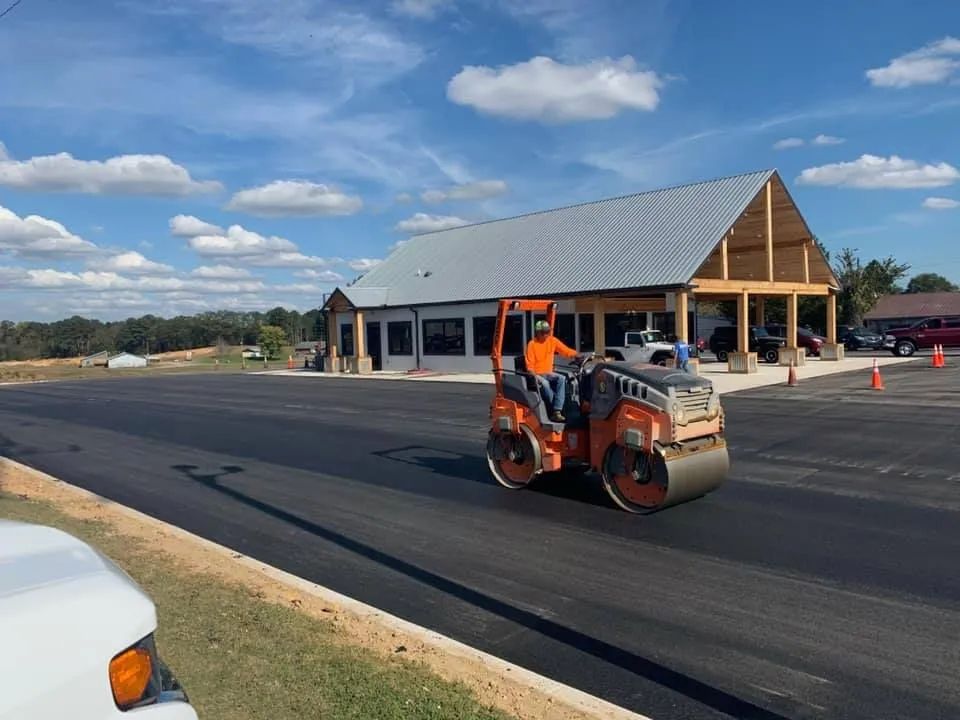 An orange steamroller drives on a freshly paved black asphalt parking lot in front of a building under a blue sky.