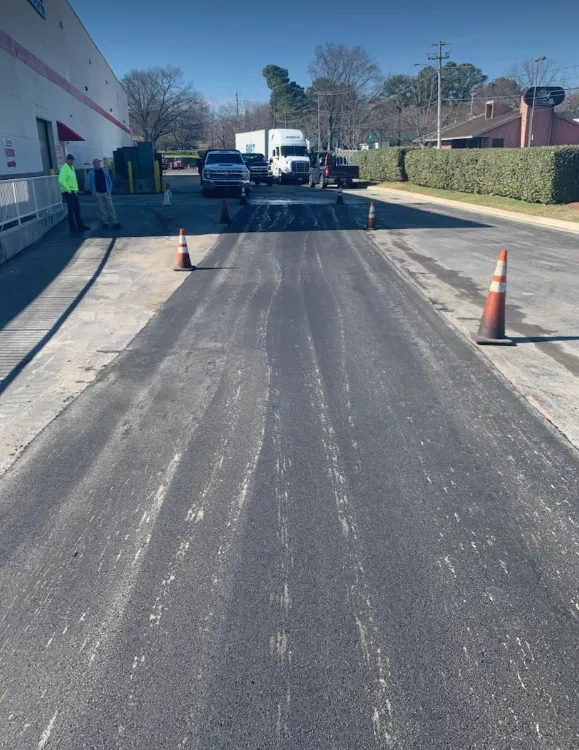 Workers stand by a building near a freshly paved section of a parking lot marked by orange traffic cones.