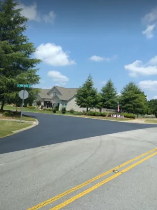 A paved road forks into a residential neighborhood with a suburban house, green trees, and a blue sky overhead.