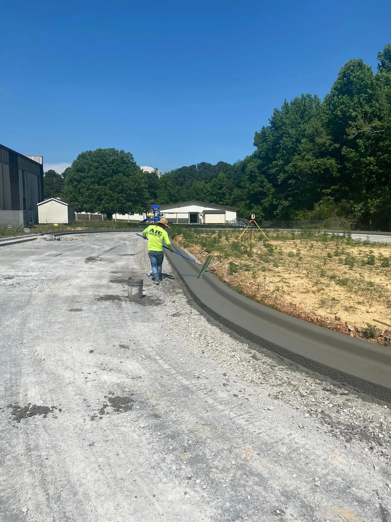 A construction worker in a high-visibility yellow vest walks beside a newly poured concrete curb on a sunny day.