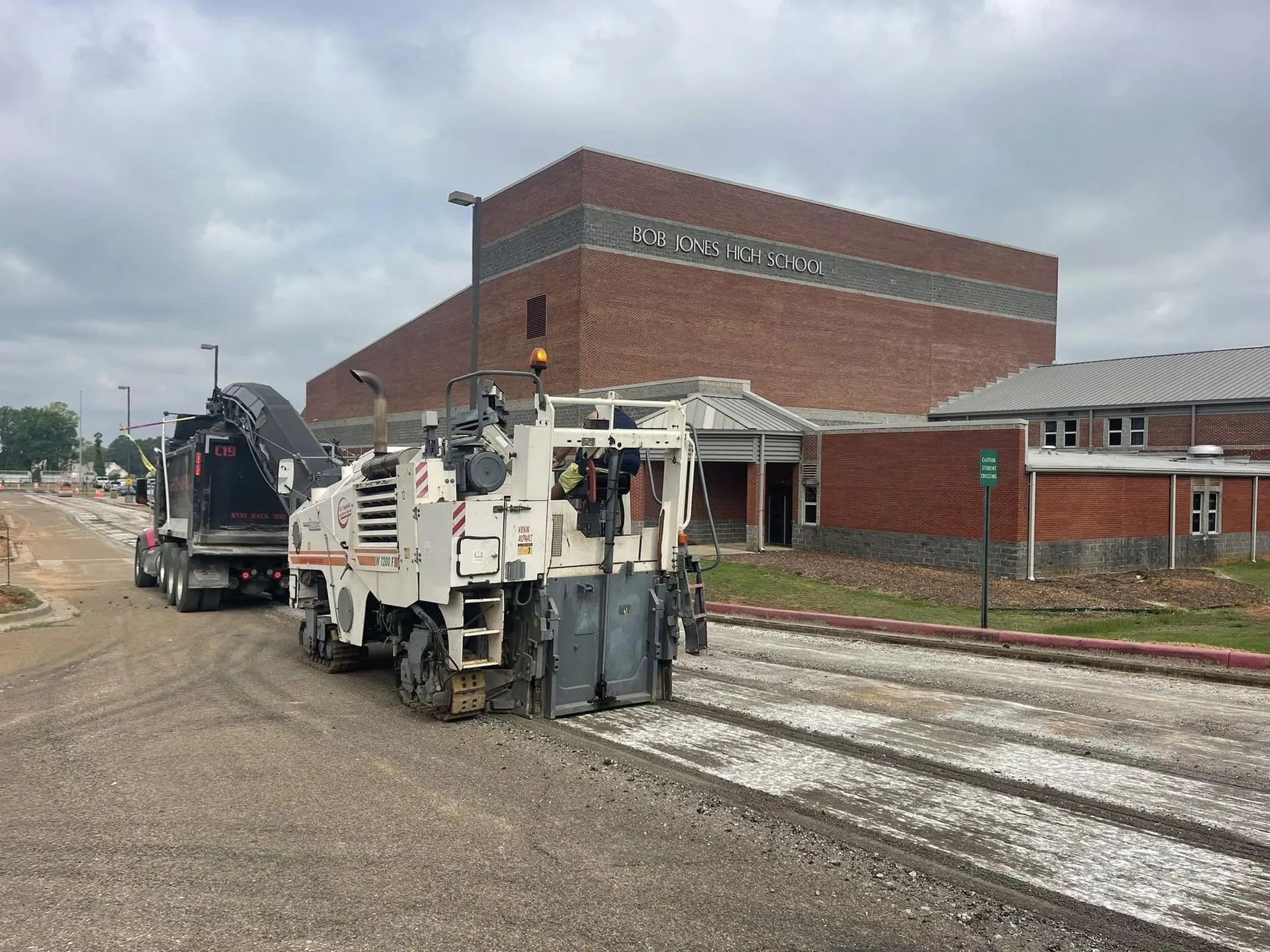 A white asphalt milling machine sits on a road near a large brick building under a cloudy sky.