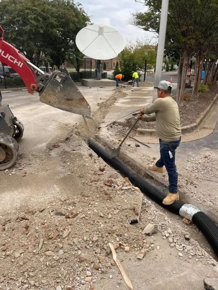 A worker uses a tool to guide a black pipe into a trench alongside a construction excavator on a paved site.