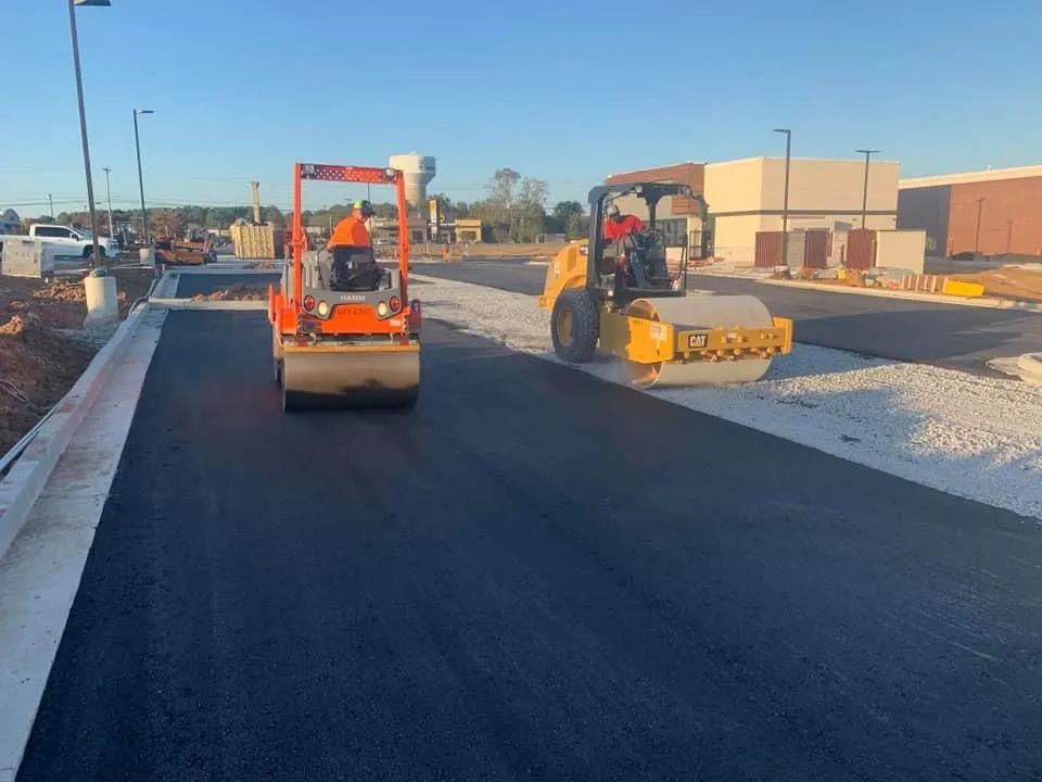 Two yellow and orange industrial rollers compacting freshly laid black asphalt on a sunny construction site.
