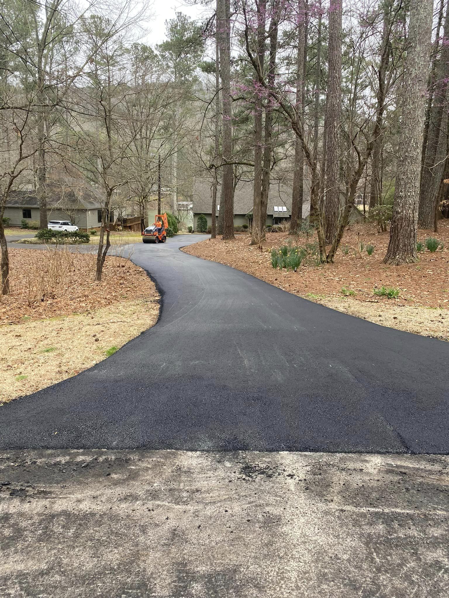 A fresh, dark asphalt driveway curves through a wooded residential yard toward a house in the distance.