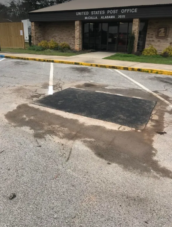 A patch of new asphalt on a gravel parking lot in front of a United States Post Office building.