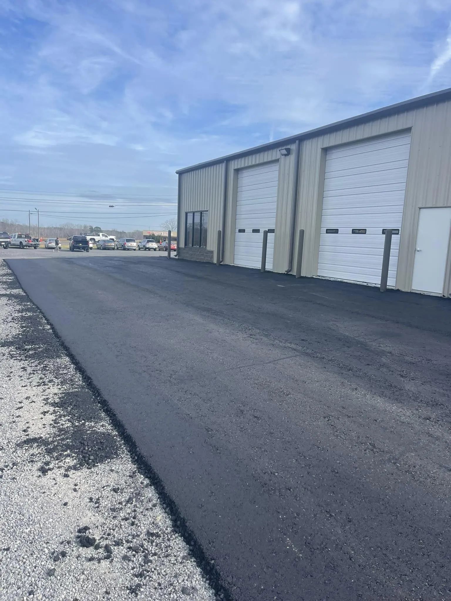 A freshly paved black asphalt driveway leading to a metal warehouse building with two white garage doors under a blue sky.
