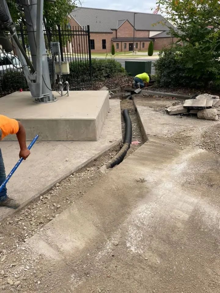 Workers excavate a trench near a concrete tower base, installing a black conduit pipe in an outdoor work site.