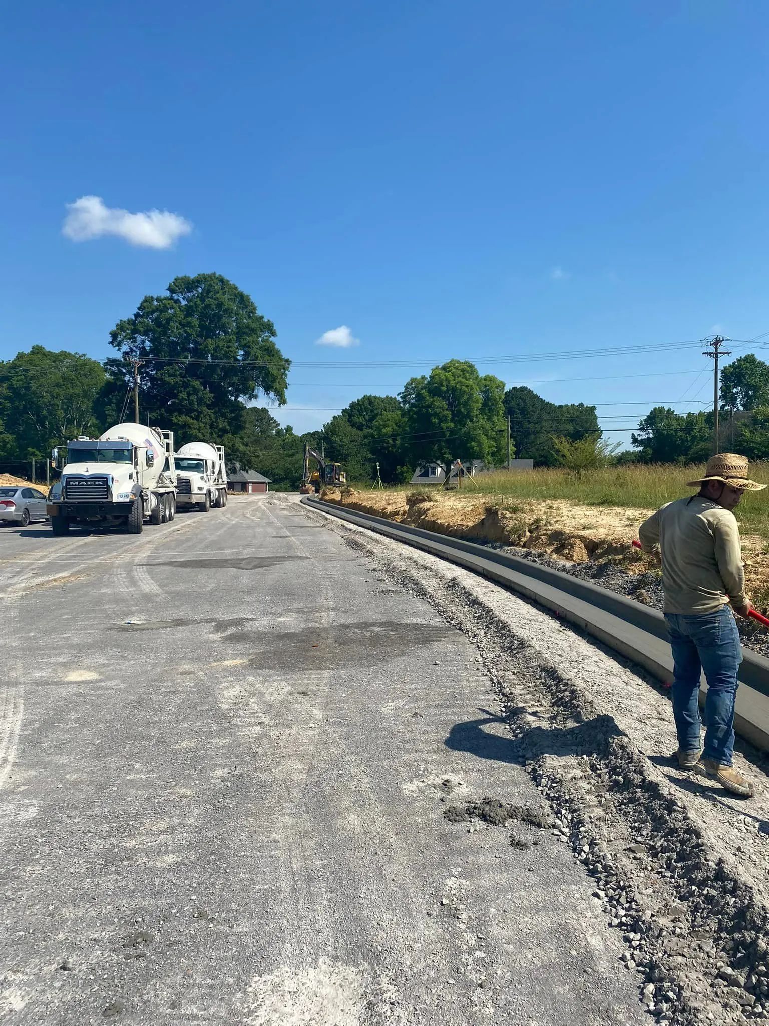 A worker oversees the installation of concrete curbing along a gravel construction site with two cement trucks nearby.