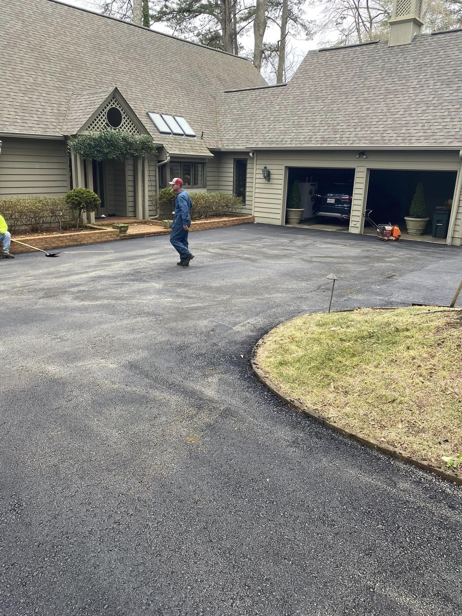 A person in blue coveralls walks across a freshly paved asphalt driveway in front of a house with a garage.