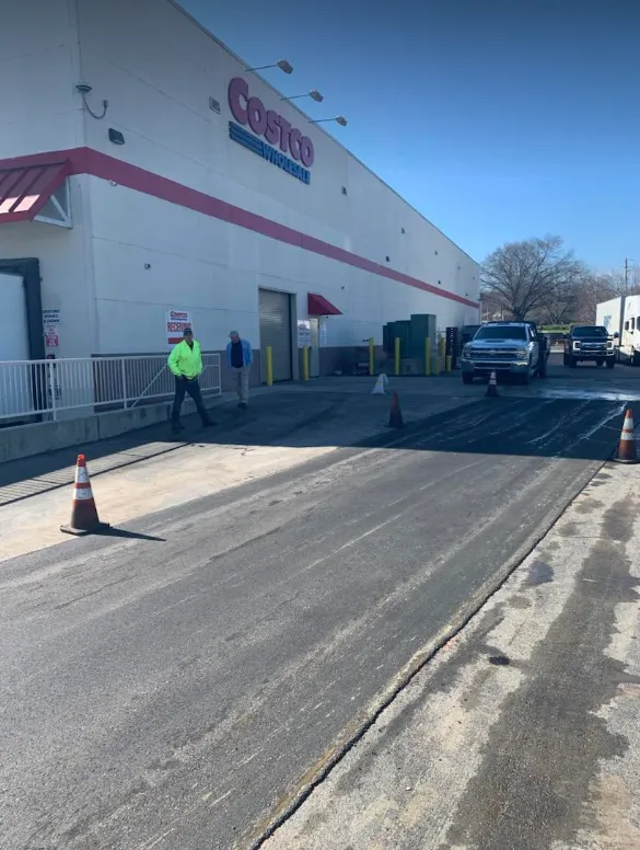 Two workers in reflective vests stand near a partially paved loading area outside a Costco Wholesale building.