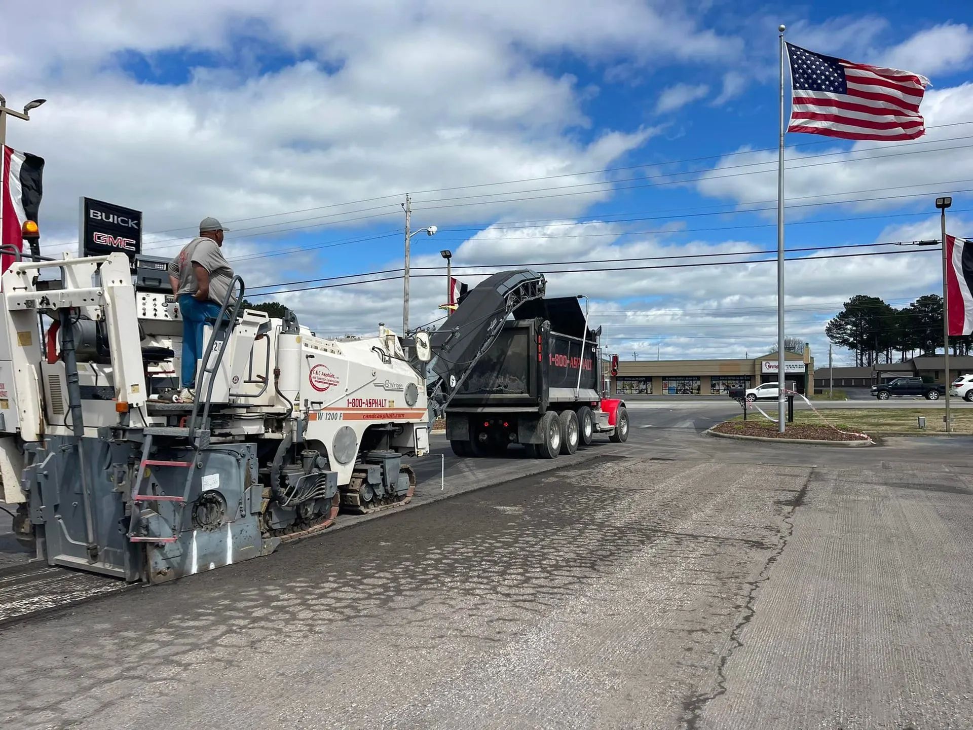 A construction crew operates a milling machine to remove asphalt into a dump truck under a bright blue sky with an US flag.