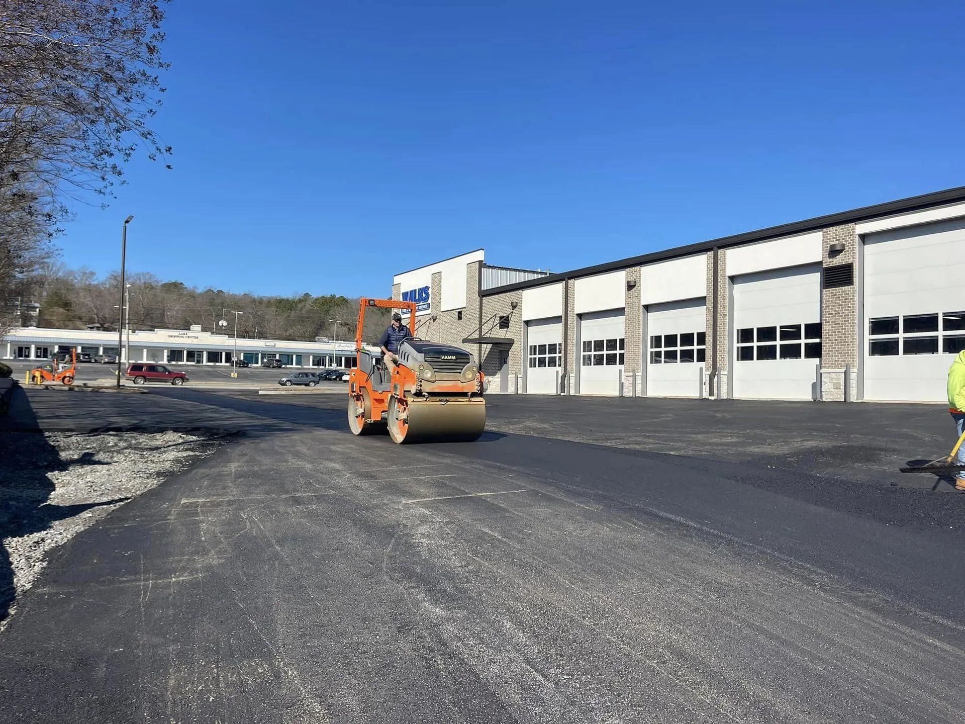 A bright orange construction roller compacts fresh asphalt in a parking lot next to a white industrial building.