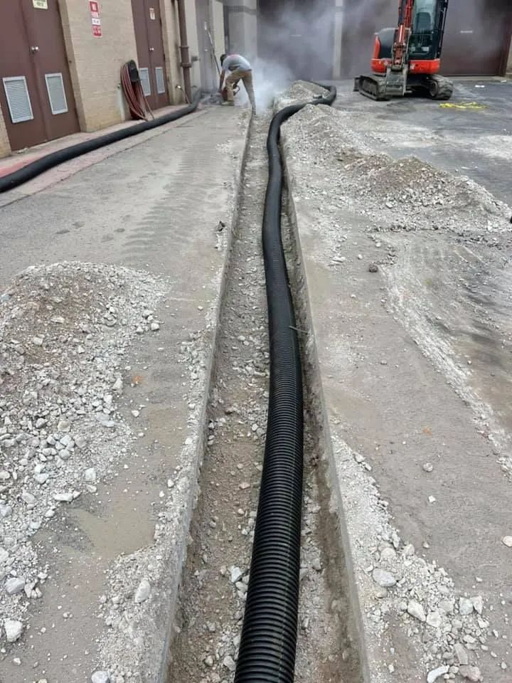 A worker cuts concrete alongside a long, black corrugated pipe laid in a narrow trench at an outdoor construction site.