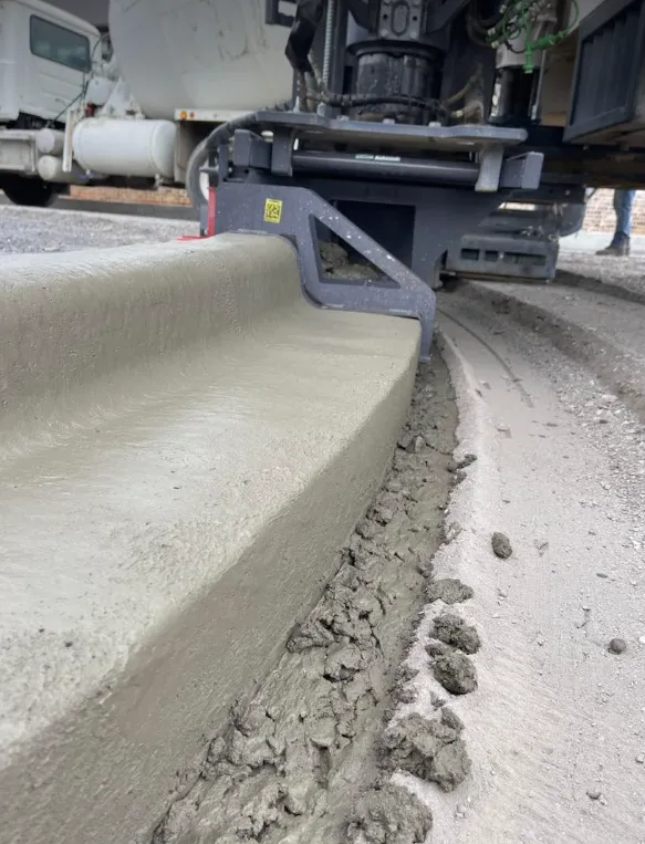 A concrete slipform machine extrudes a new curb along a gravel road, with fresh, grey concrete forming the curb profile.