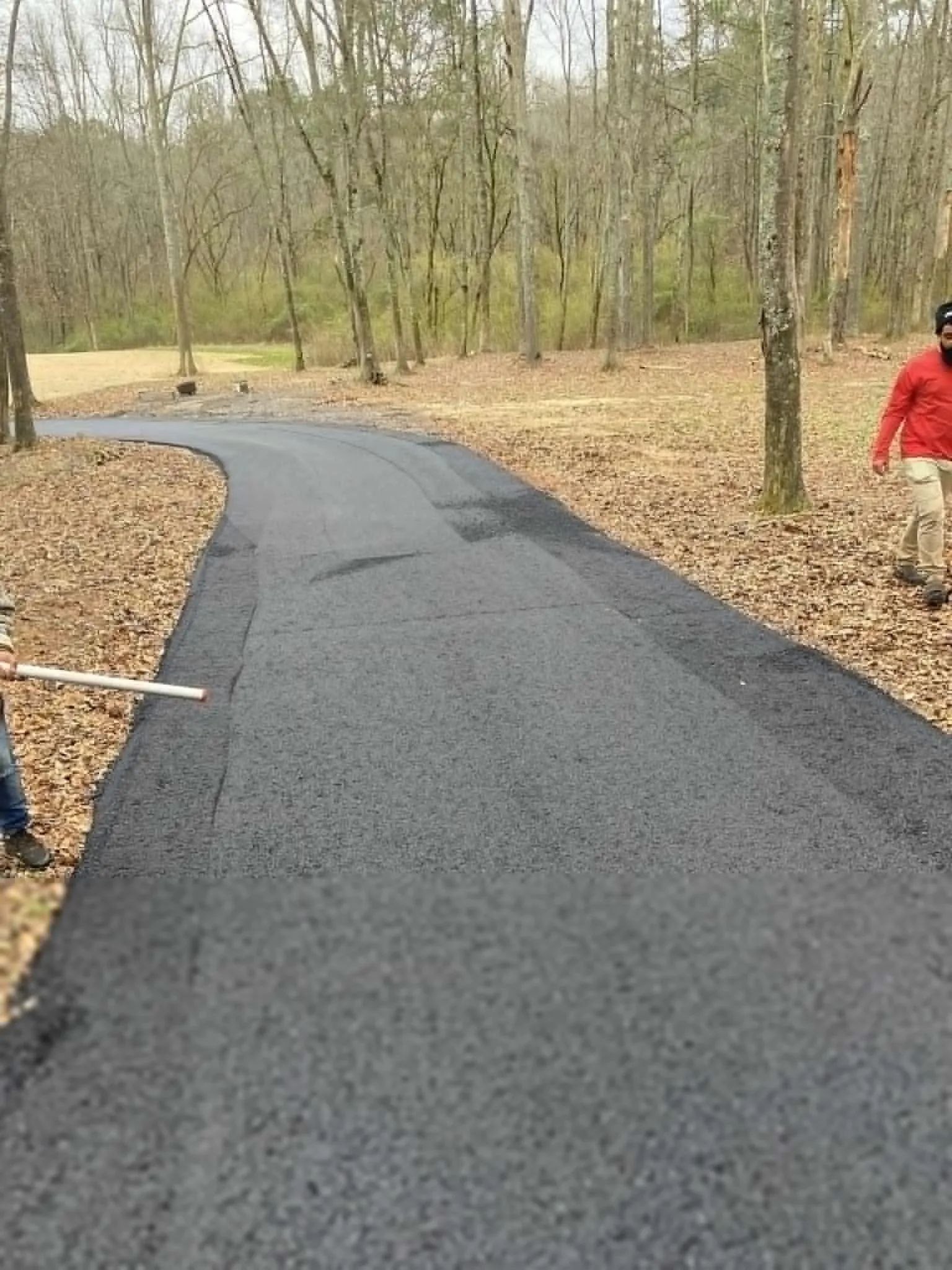 A freshly paved black asphalt driveway winds through a wooded area with fallen leaves on the ground.