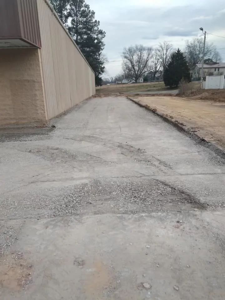 A cracked, uneven concrete driveway next to a long beige building, leading toward an open field under a cloudy sky.