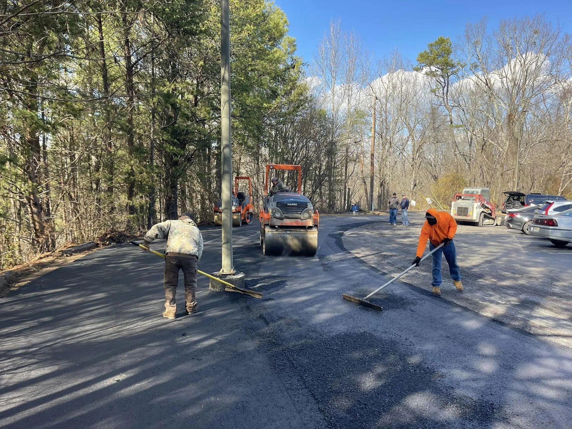 Two workers in reflective vests use rakes to spread fresh asphalt on a road while a heavy steamroller operates nearby.