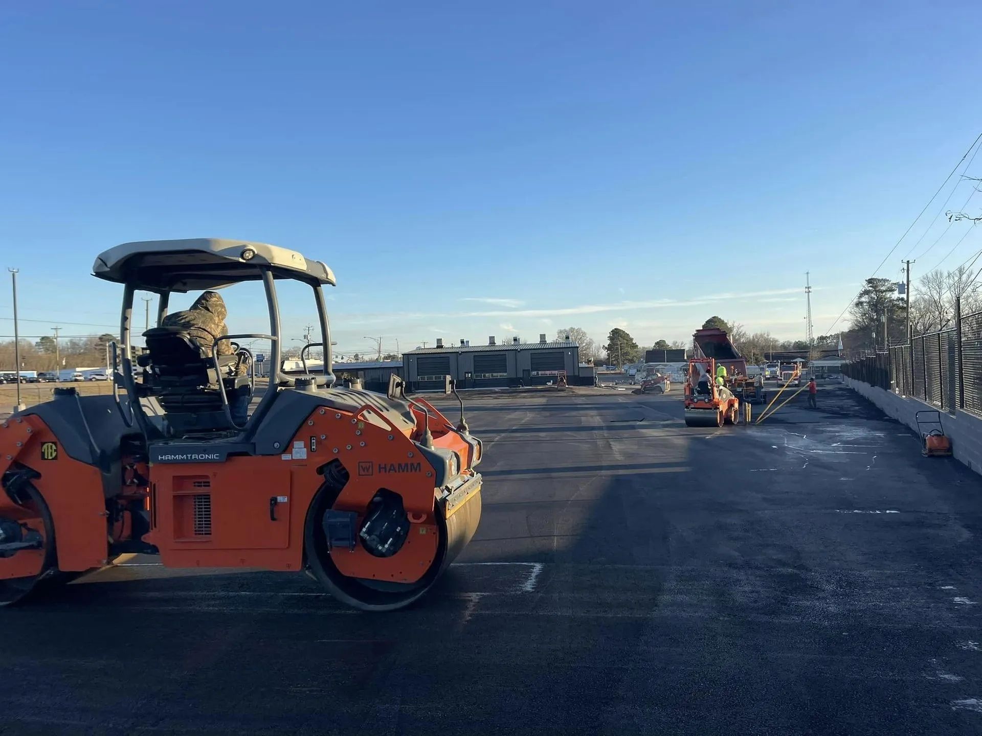 An orange construction roller on a paved lot under a clear blue sky, with a worker in the distance.