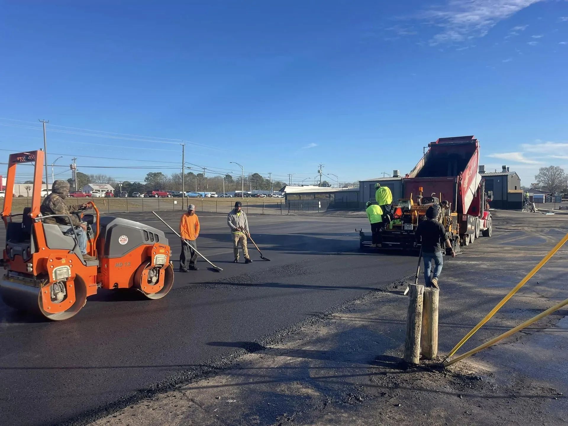 Workers pave an outdoor lot with an orange asphalt roller and a dump truck under a bright blue sky.