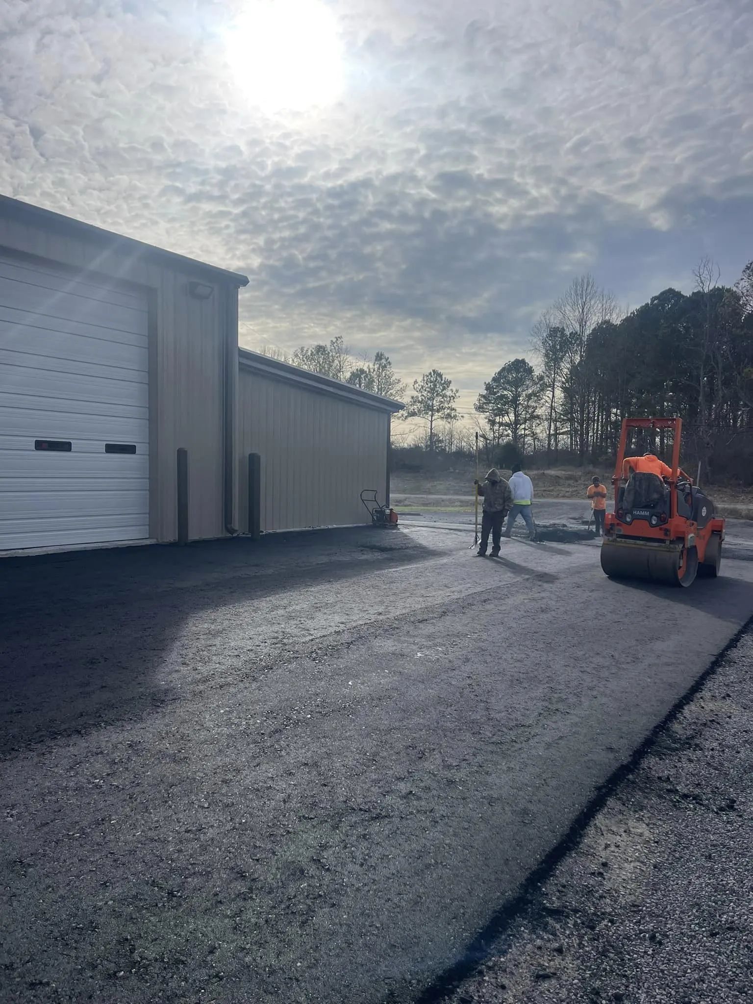 Workers use a steamroller to pave fresh asphalt in front of a tan metal warehouse building under a sunny sky.