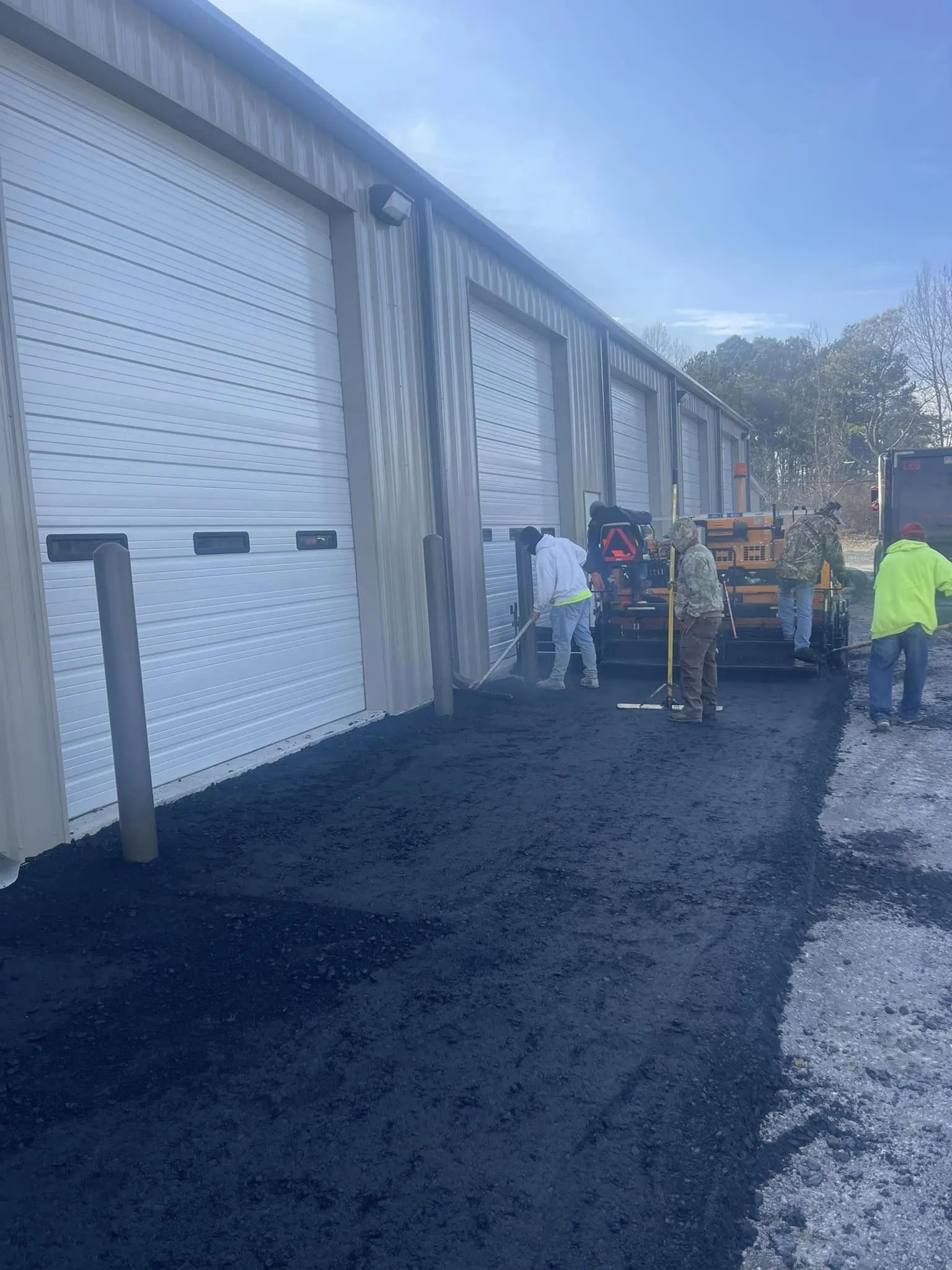 Workers in safety vests paving a fresh asphalt driveway in front of industrial warehouse roll-up doors.