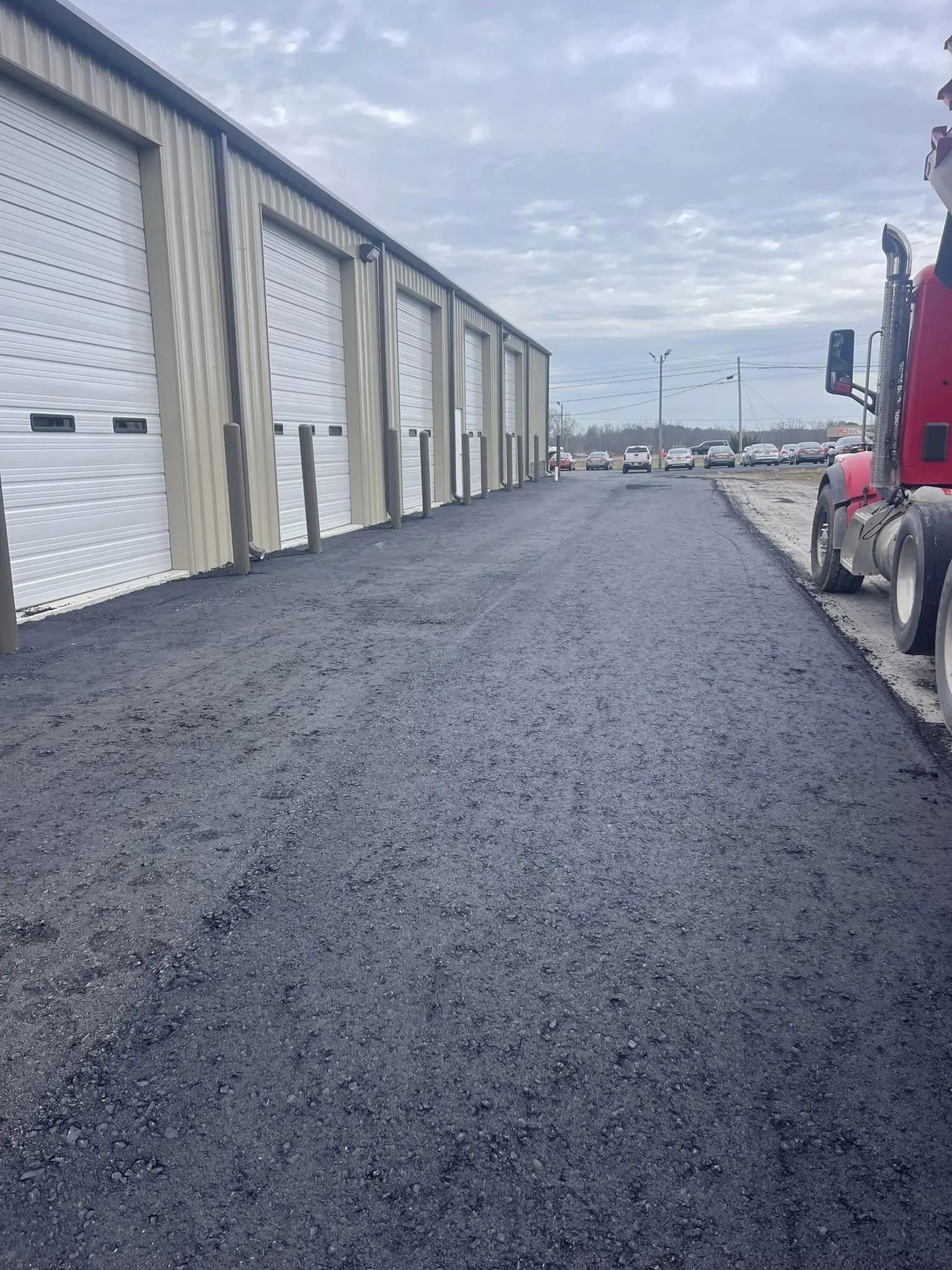 A row of storage units with white roll-up doors beside a newly paved, dark asphalt driveway and a parked red semi-truck.