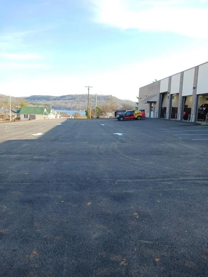 A large asphalt parking lot with a multi-bay industrial building on the right and a distant view of hills and a lake.