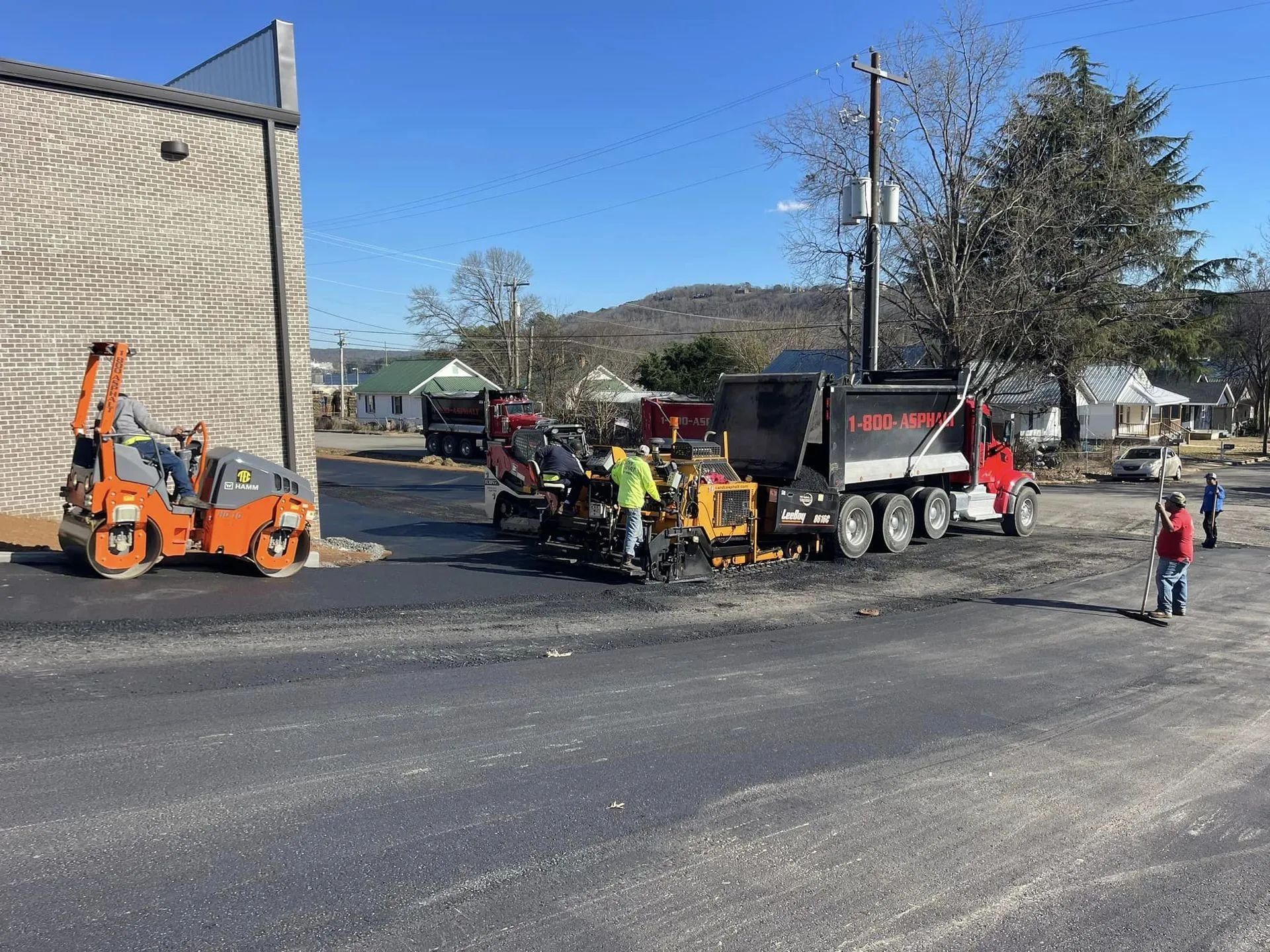 Construction workers operate an orange asphalt roller and paving machinery on a sunny day in a paved lot.