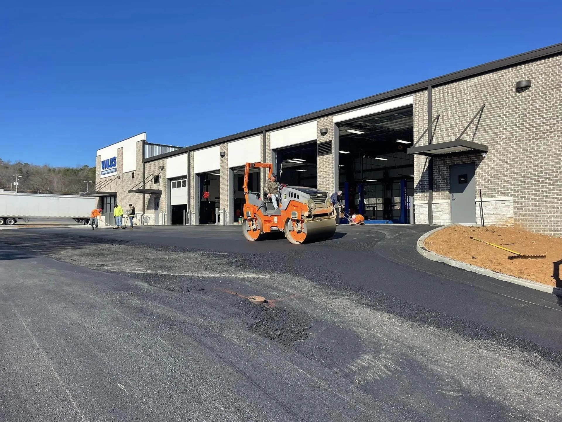 An orange construction roller moves across a newly paved asphalt lot in front of a commercial garage.