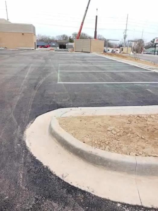 A freshly paved parking lot featuring painted white parking stalls, a curved concrete curb, and an unlandscaped dirt patch.