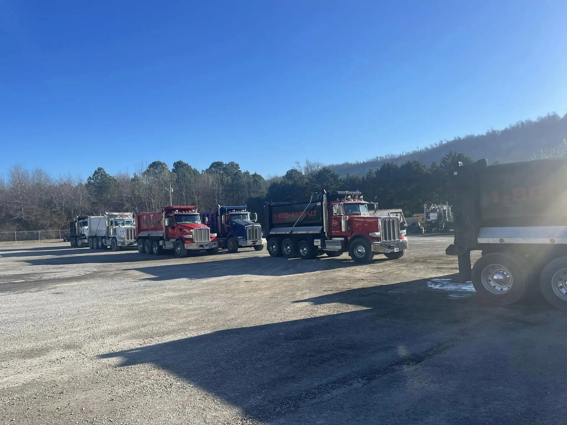 A line of red and white dump trucks parked on a gravel lot under a clear blue sky.