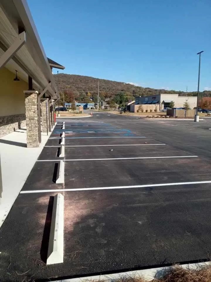 A row of parking spaces with concrete wheel stops next to a building with stone pillars under a clear blue sky.