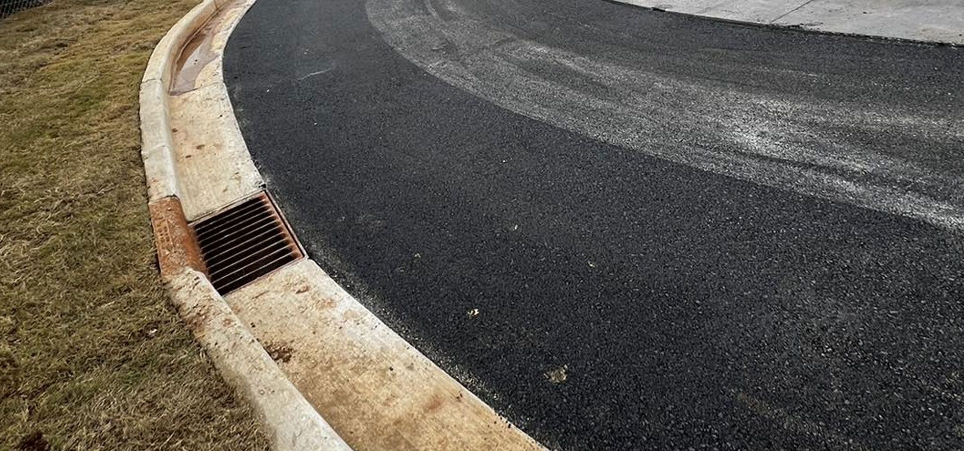 A newly paved asphalt road curves along a concrete curb with a metal storm drain grate set into the gutter.