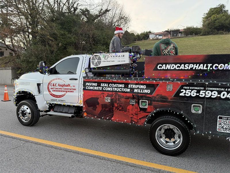 A white utility truck for C and C Asphalt, decorated with holiday lights, drives down a road with two people in the bed.