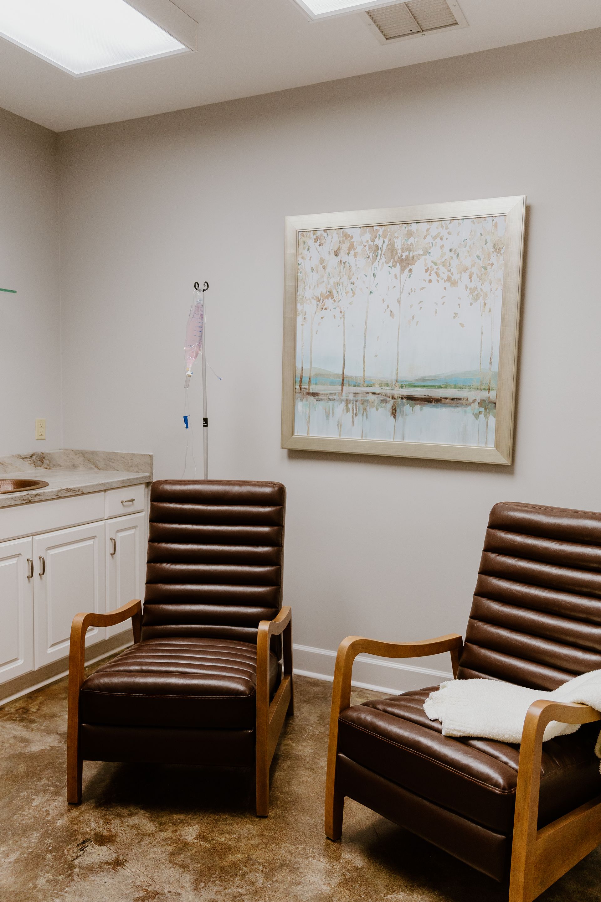 Two brown leather reclining chairs placed in a waiting or clinical room with white cabinets and a framed landscape painting.
